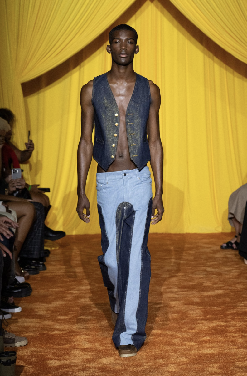 Slender male model with deep skin wearing an unbuttoned denim vest and pants walking on a runway against a yellow backdrop